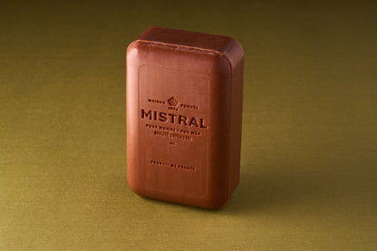 A rectangular bar of Mistral For Men Mahogany Rum Bar Soap stands upright on a plain olive-green surface. Its packaging features embossed French text, a decorative emblem, and hints at its exclusive mahogany rum fragrance.