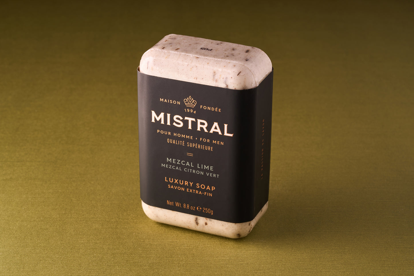 A Mistral For Men Mezcal Lime Bar Soap, triple-milled and rectangular, rests on a greenish-brown surface. Its black wrapper with gold and white text suggests a French fragrance, while visible specks enhance its luxurious look.