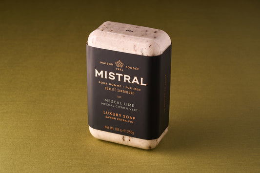 A Mistral For Men Mezcal Lime Bar Soap, triple-milled and rectangular, rests on a greenish-brown surface. Its black wrapper with gold and white text suggests a French fragrance, while visible specks enhance its luxurious look.