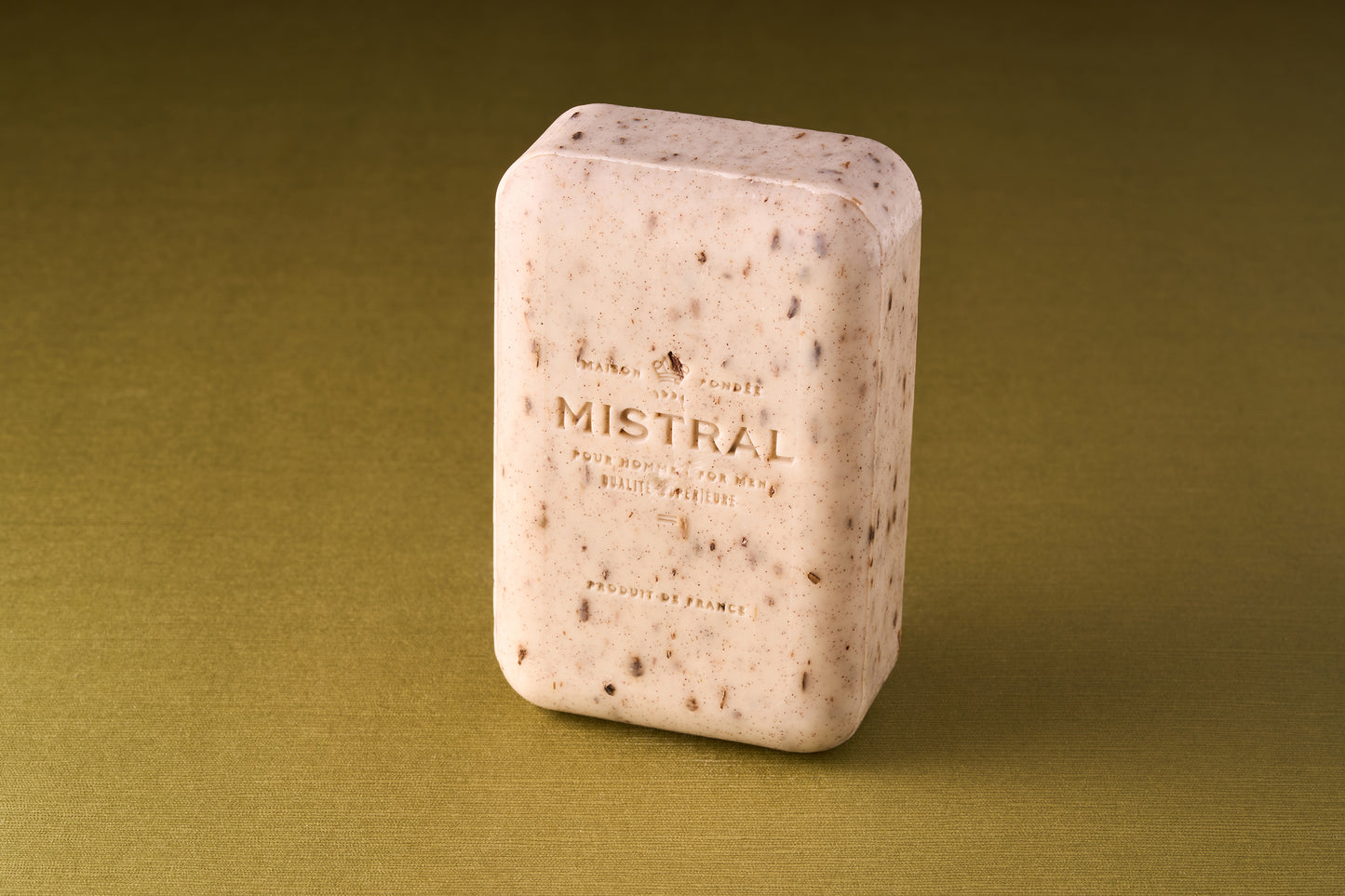 A rectangular bar of Mistral For Men Mezcal Lime Bar Soap with visible exfoliating particles stands upright on an olive-green surface. The light beige, triple-milled soap has its name embossed, highlighting its premium quality.