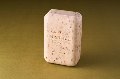 A rectangular bar of Mistral For Men Mezcal Lime Bar Soap with visible exfoliating particles stands upright on an olive-green surface. The light beige, triple-milled soap has its name embossed, highlighting its premium quality.