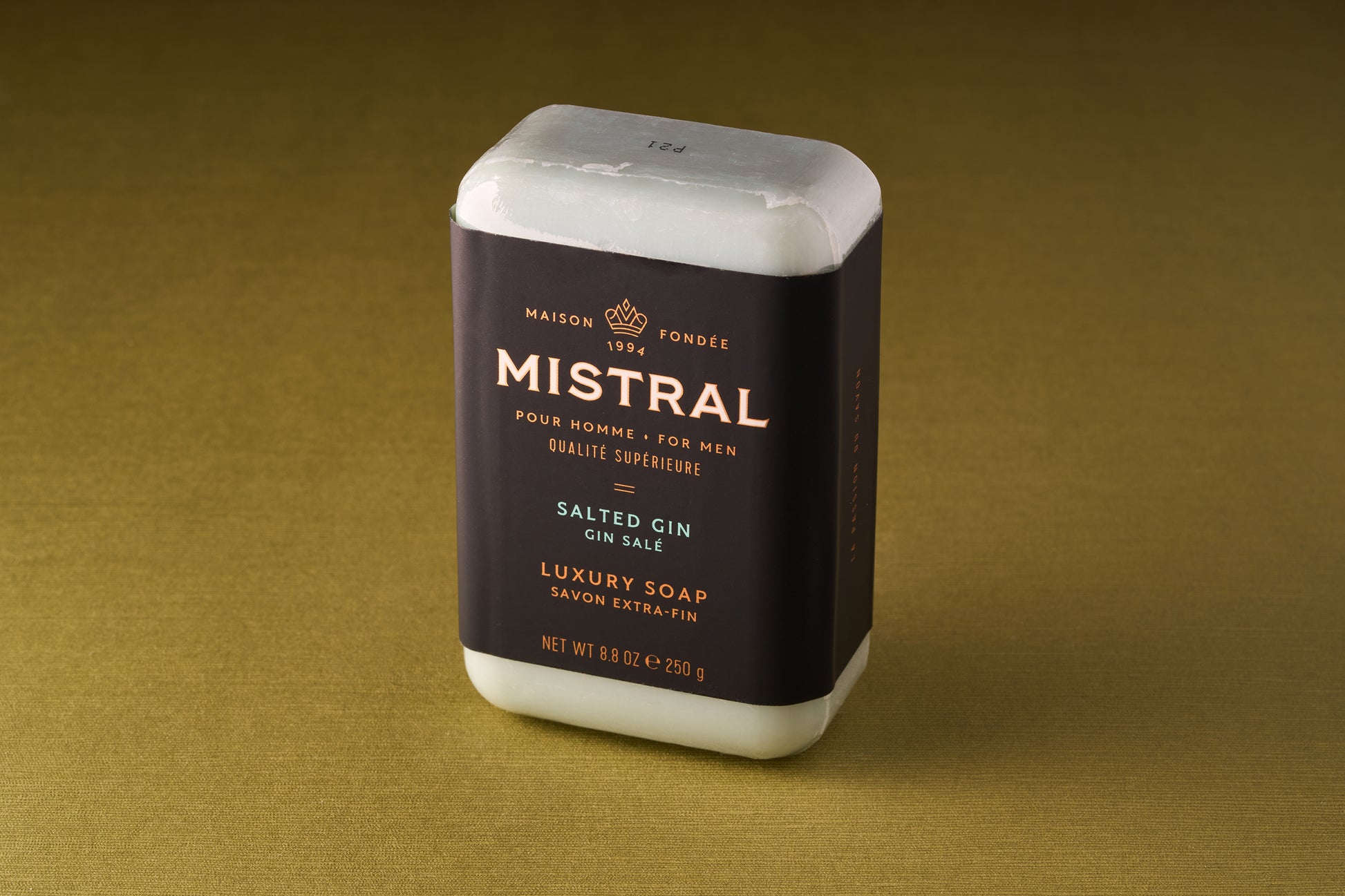 An 8 oz (250g) bar of Mistral For Men Salted Gin Bar Soap with a black and gold label stands upright on a gold surface. This triple-milled luxury soap features a refined French fragrance crafted for men.