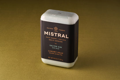 An 8 oz (250g) bar of Mistral For Men Salted Gin Bar Soap with a black and gold label stands upright on a gold surface. This triple-milled luxury soap features a refined French fragrance crafted for men.