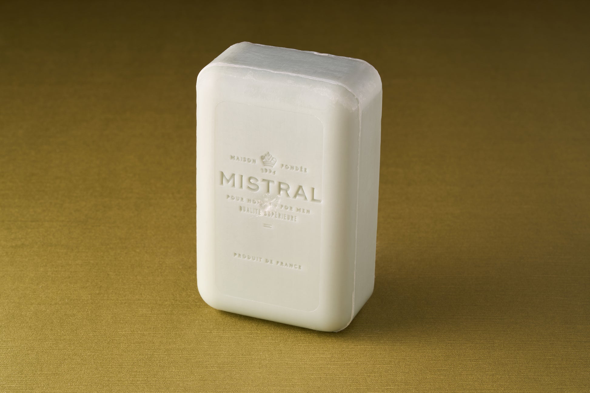 A rectangular white bar of Mistral For Men Salted Gin Bar Soap stands upright on a textured golden-brown surface. Embossed text and a minimalist design showcase its refined French fragrance for men.