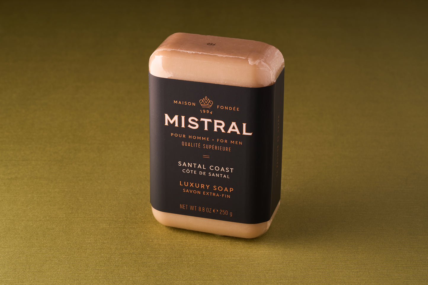 A Mistral For Men Santal Coast Bar Soap, infused with French fragrance and wrapped in black and gold packaging, rests on a plain olive-green surface.
