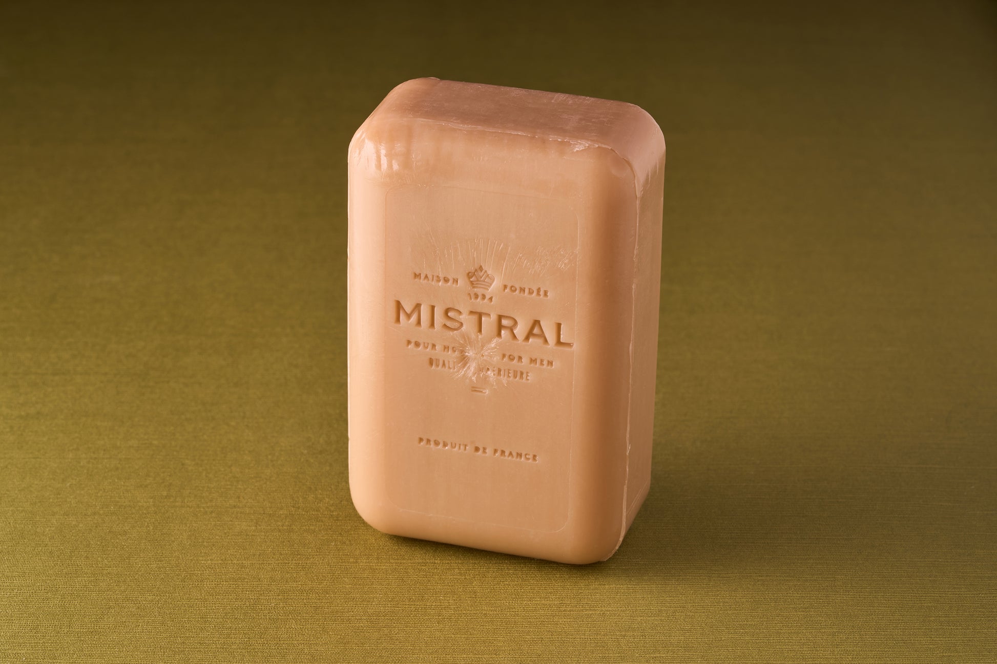 A rectangular Mistral For Men Santal Coast Bar Soap stands upright on a green surface, featuring embossed text and logo on its peach-colored front, reflecting the sophistication of Provence-inspired French fragrance.