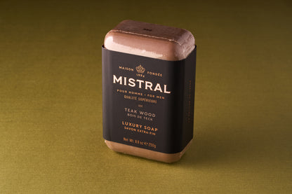 The Mistral For Men Teak Wood Bar Soap, in black and gold packaging, rests on an olive green surface.