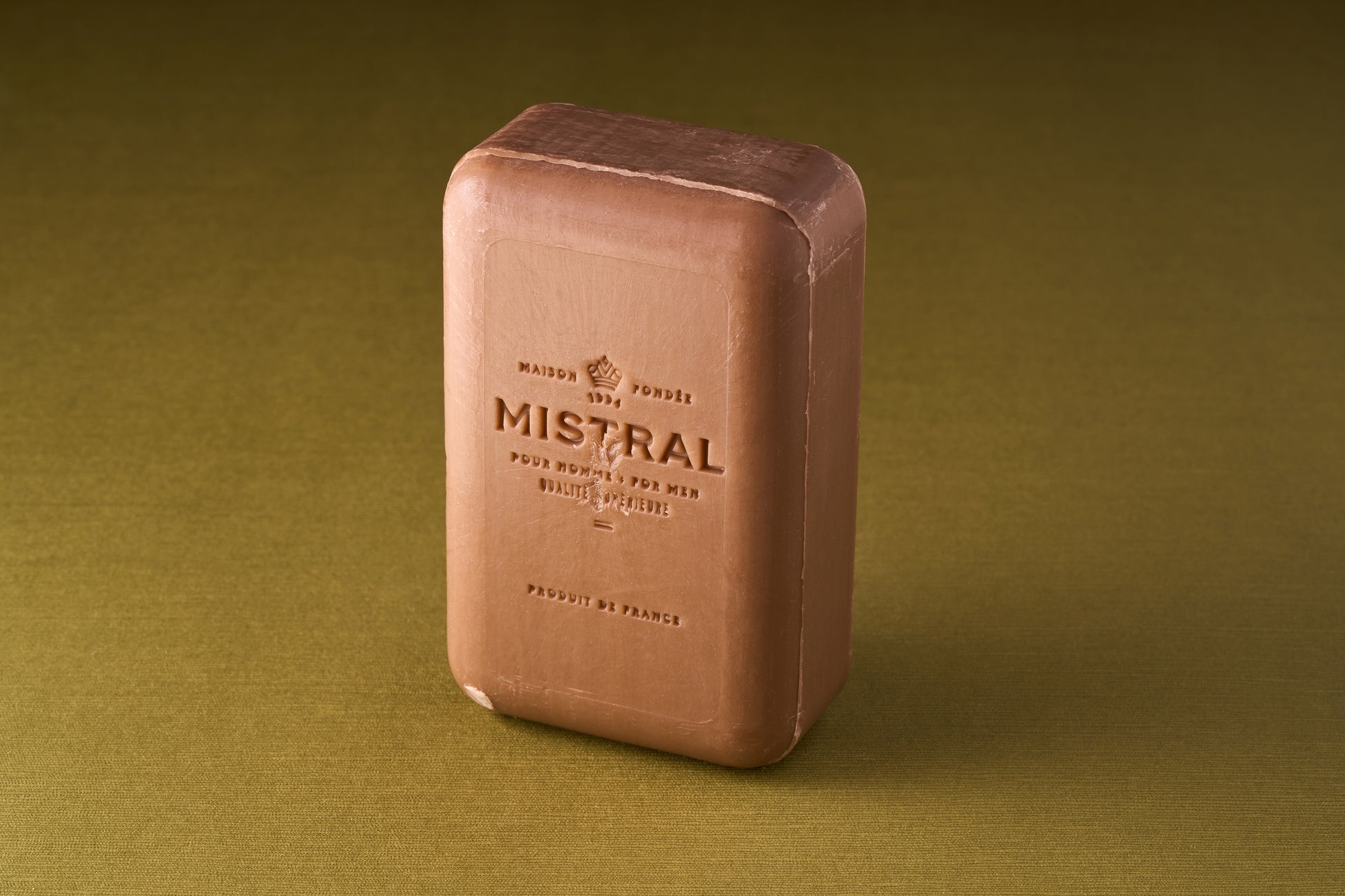 A rectangular Mistral For Men Teak Wood Bar Soap stands upright on a green textured surface. The triple-milled soap features embossed text and a logo, adding to its classic, artisanal appeal.