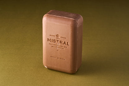A rectangular Mistral For Men Teak Wood Bar Soap stands upright on a green textured surface. The triple-milled soap features embossed text and a logo, adding to its classic, artisanal appeal.