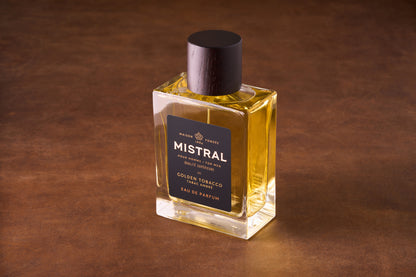 A 100ml bottle of Mistral Golden Tobacco Eau de Parfum, featuring a black label with gold and white text and topped with a dark cap, contains yellow liquid and rests on a brown surface.