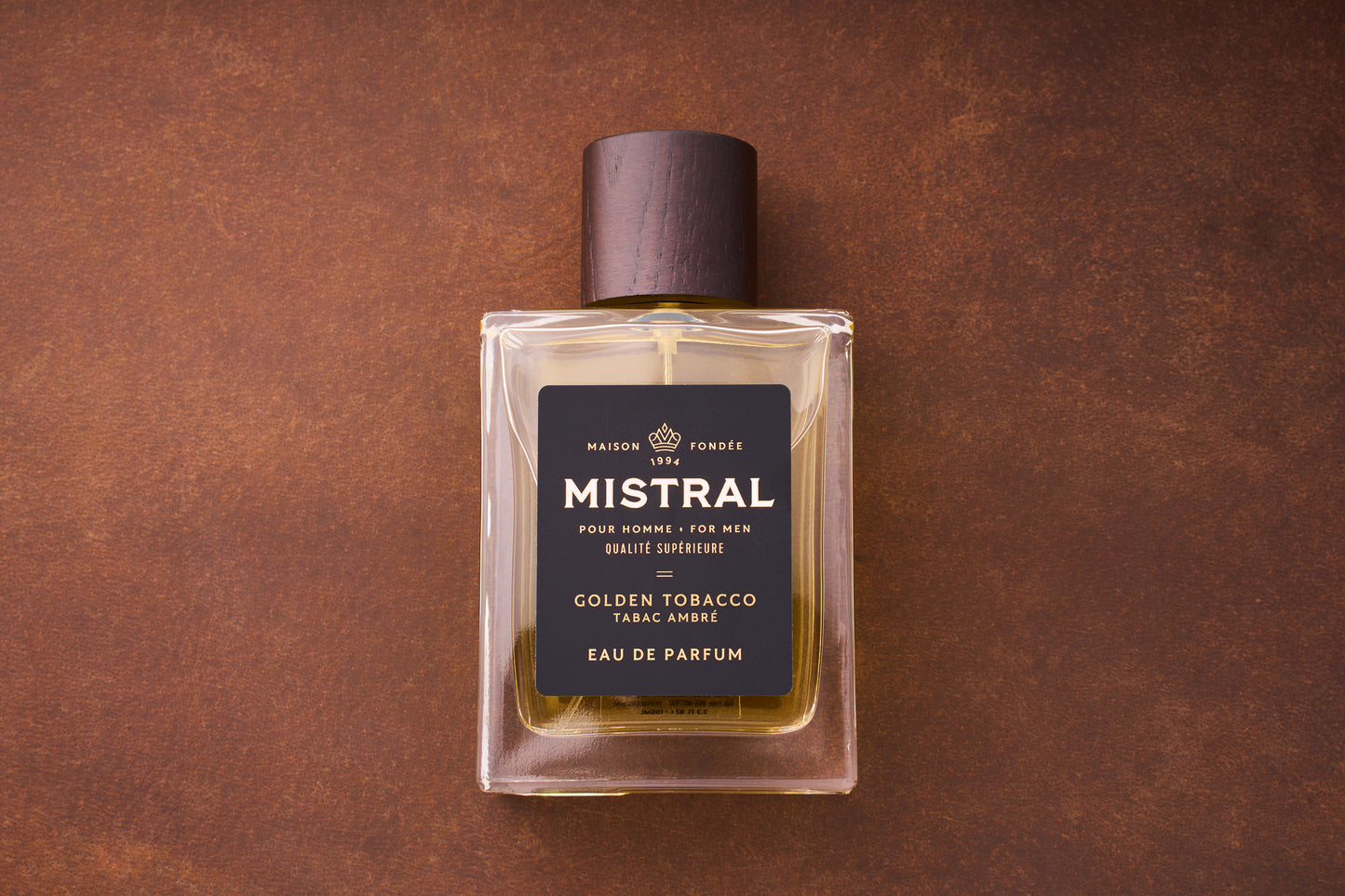 A 100ml bottle of Mistral Golden Tobacco Eau de Parfum, featuring a dark brown cap, gold-hued liquid, and a black and gold label, sits on a brown surface.