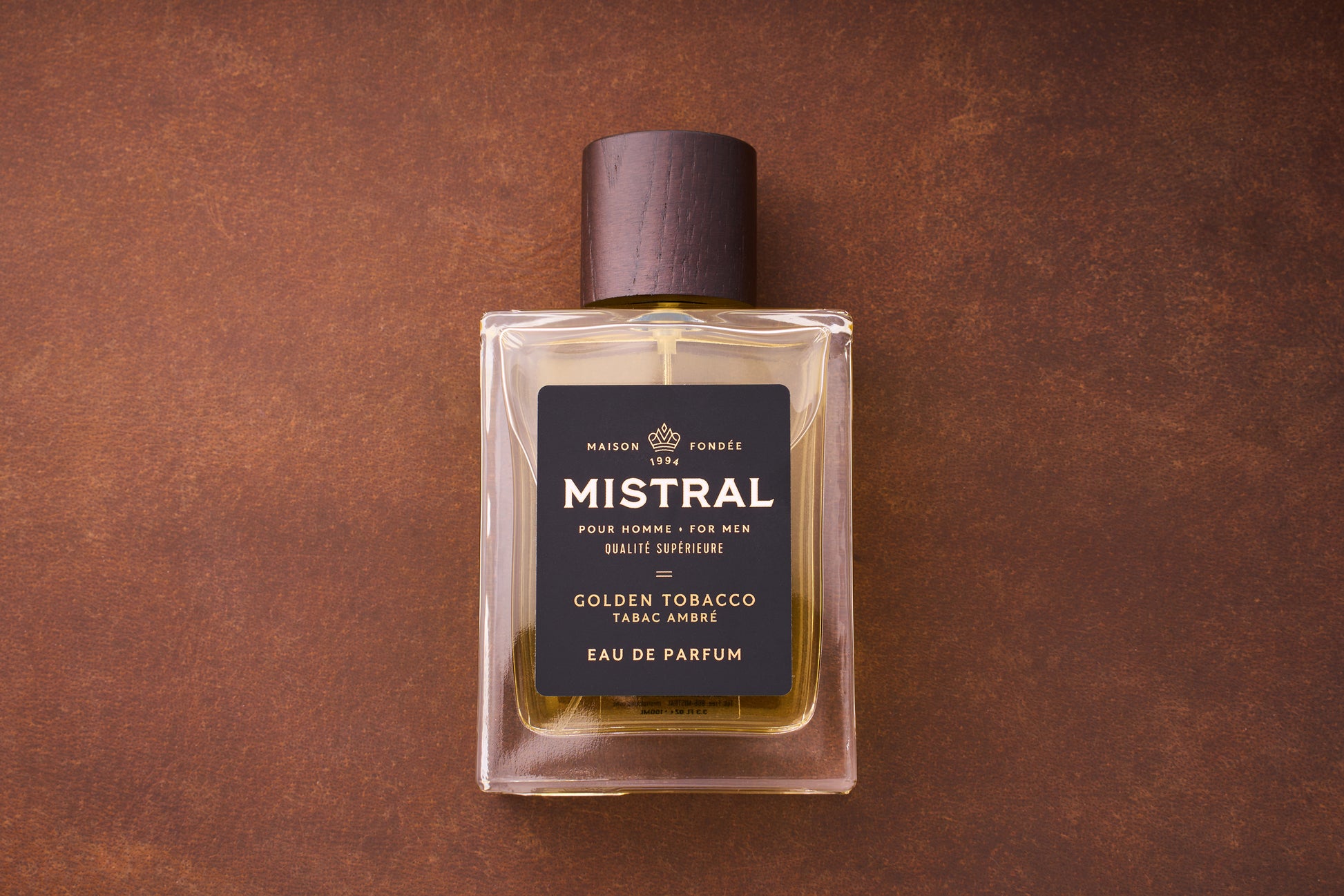 A 100ml bottle of Mistral Golden Tobacco Eau de Parfum, featuring a dark brown cap, gold-hued liquid, and a black and gold label, sits on a brown surface.