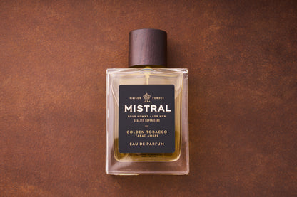 A 100ml bottle of Mistral Golden Tobacco Eau de Parfum, featuring a dark brown cap, gold-hued liquid, and a black and gold label, sits on a brown surface.