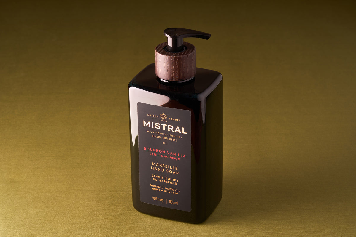 A dark rectangular pump bottle of Mistral Liquid Hand Soap - Bourbon Vanilla rests on a plain olive background. The label displays gold and red text, French fragrance notes, product details, and a wooden-style top.