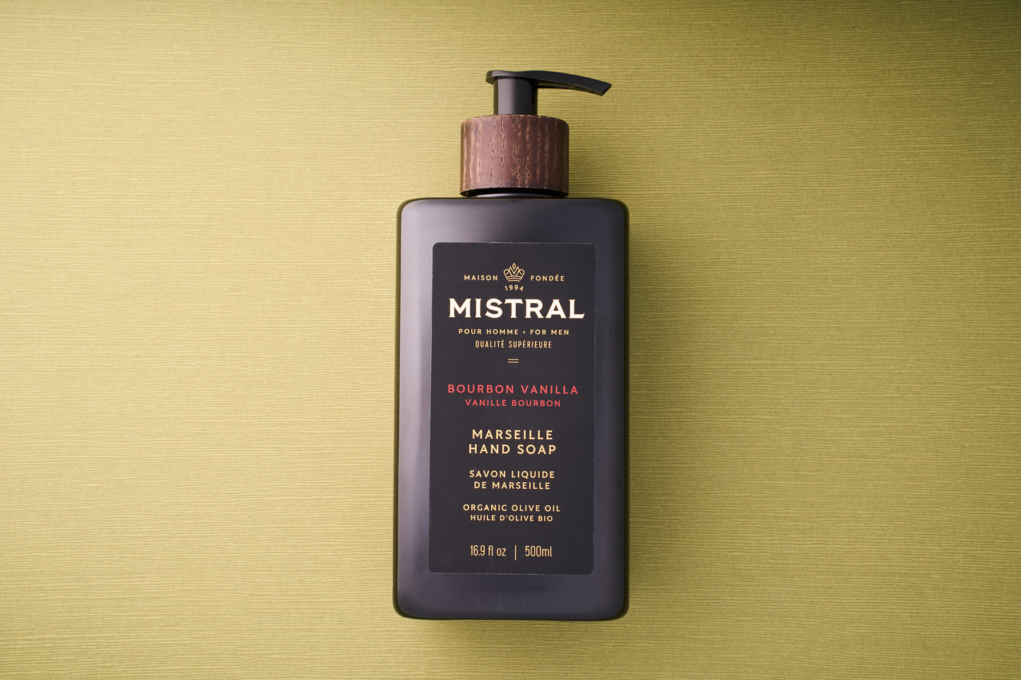 A black pump bottle of Mistral Liquid Hand Soap - Bourbon Vanilla rests on a textured green surface. The label, featuring gold and red text, notes its French fragrance and inclusion of organic olive oil.