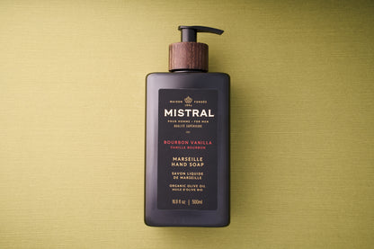 A black pump bottle of Mistral Liquid Hand Soap - Bourbon Vanilla rests on a textured green surface. The label, featuring gold and red text, notes its French fragrance and inclusion of organic olive oil.