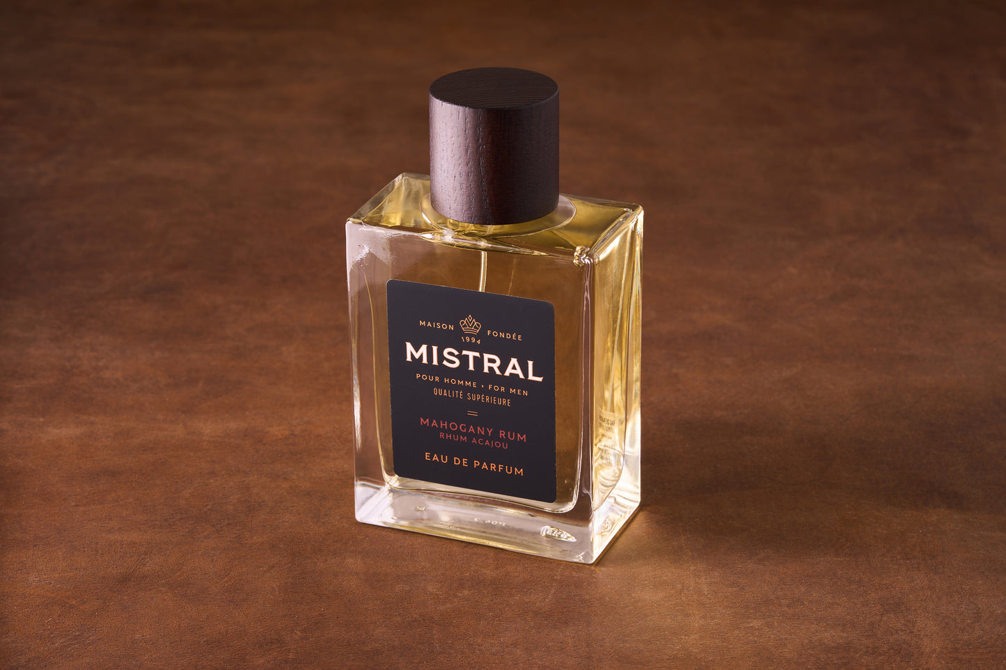 A 100ml bottle of Mistral Mahogany Rum Eau de Parfum rests on a brown textured surface, featuring a dark wood cap and gold label that emphasize its refined French craftsmanship.