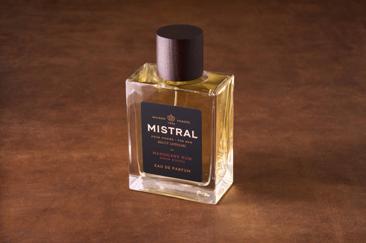 A 100ml bottle of Mistral Mahogany Rum Eau de Parfum rests on a brown textured surface, featuring a dark wood cap and gold label that emphasize its refined French craftsmanship.