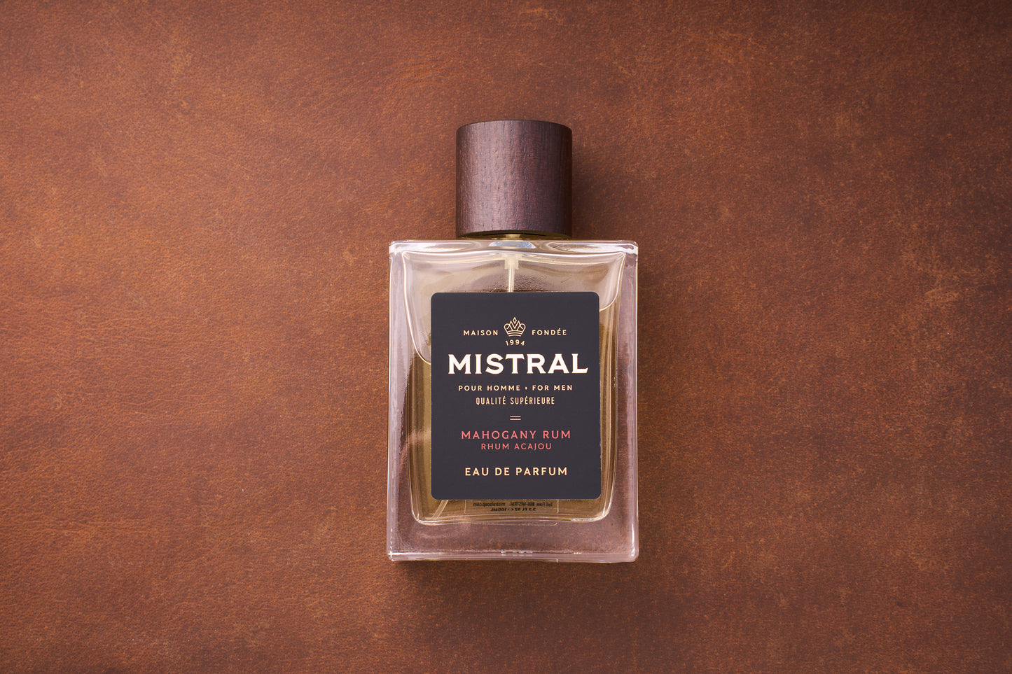 A 100ml bottle of Mistral Mahogany Rum Eau de Parfum for men, inspired by Provence, features a dark wooden cap on a brown textured surface. Its black label displays elegant gold and white text.