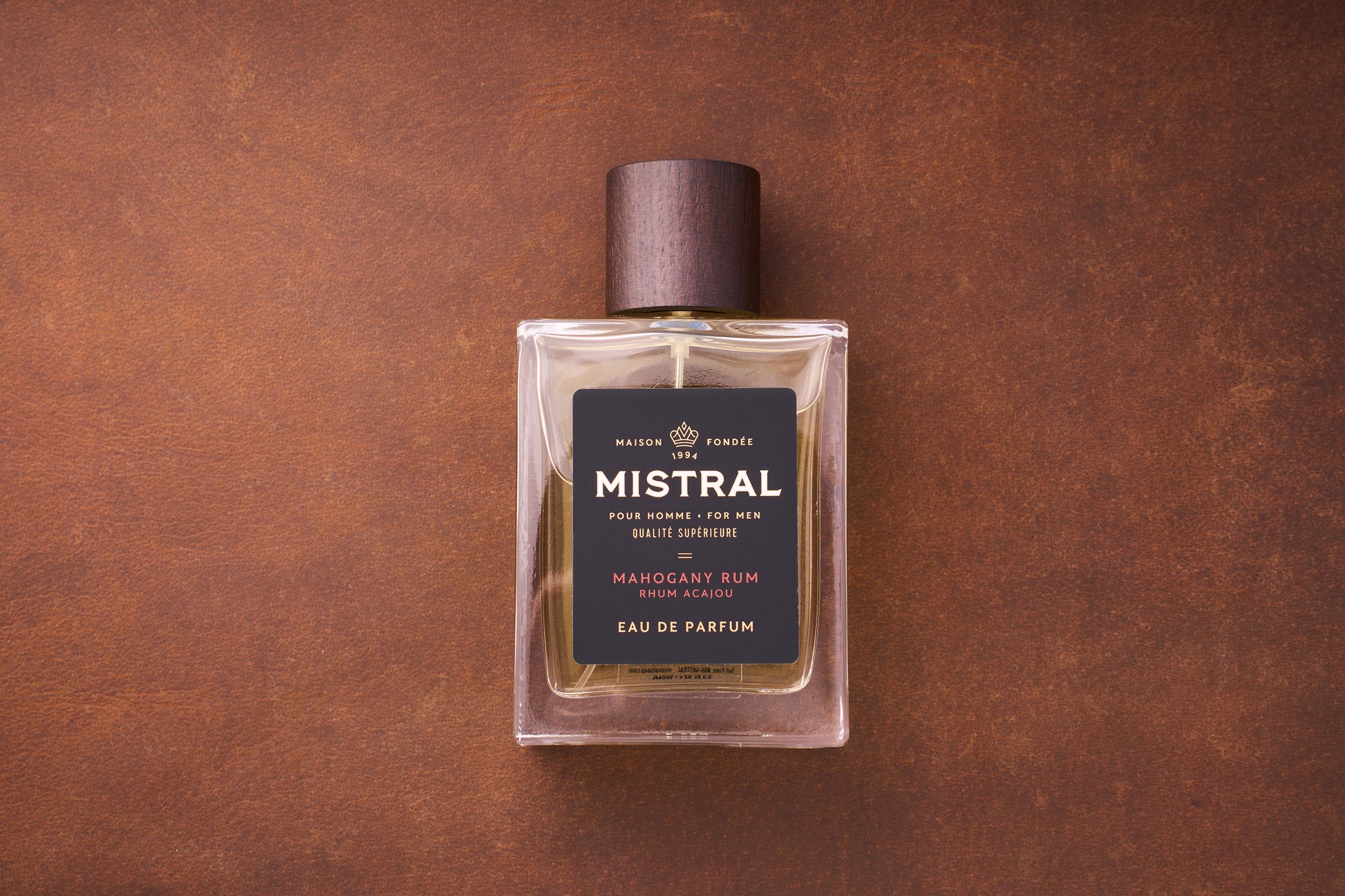 A 100ml bottle of Mistral Mahogany Rum Eau de Parfum for men, inspired by Provence, features a dark wooden cap on a brown textured surface. Its black label displays elegant gold and white text.