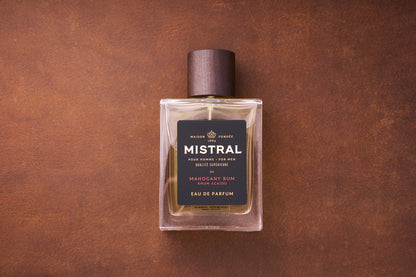 A 100ml bottle of Mistral Mahogany Rum Eau de Parfum for men, inspired by Provence, features a dark wooden cap on a brown textured surface. Its black label displays elegant gold and white text.