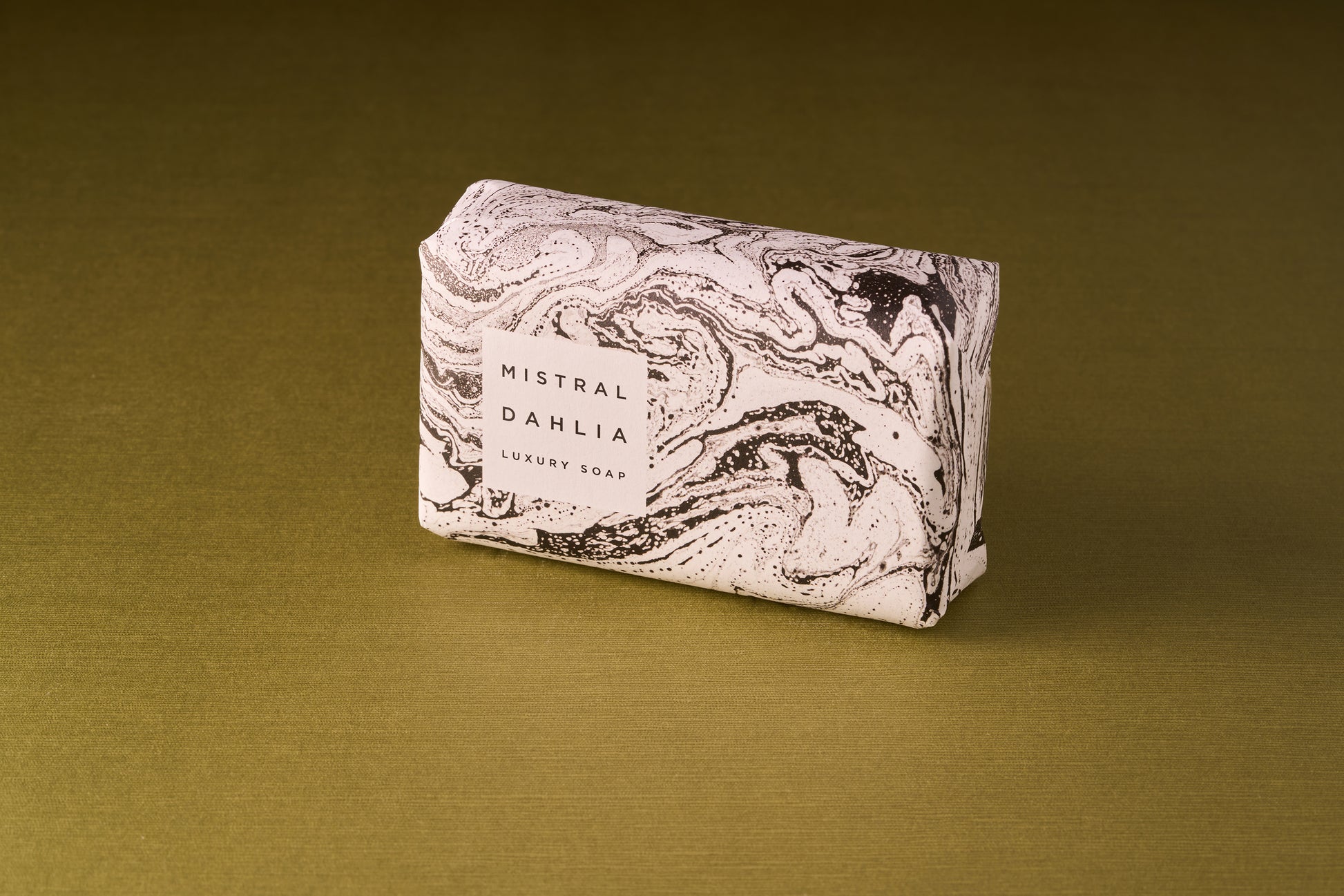 A Mistral Marbles Dahlia Bar Soap, wrapped in white paper with black marble patterns, is displayed on a gold surface.