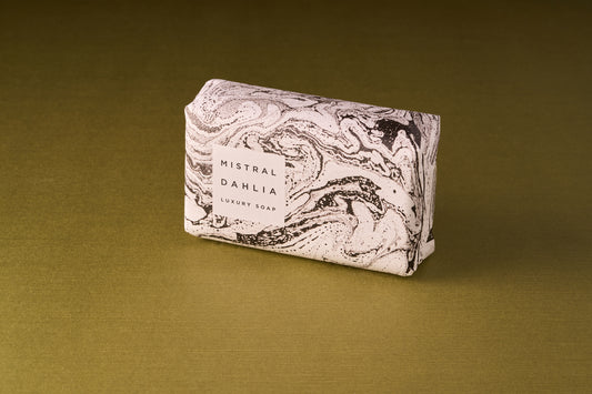 A Mistral Marbles Dahlia Bar Soap, wrapped in white paper with black marble patterns, is displayed on a gold surface.