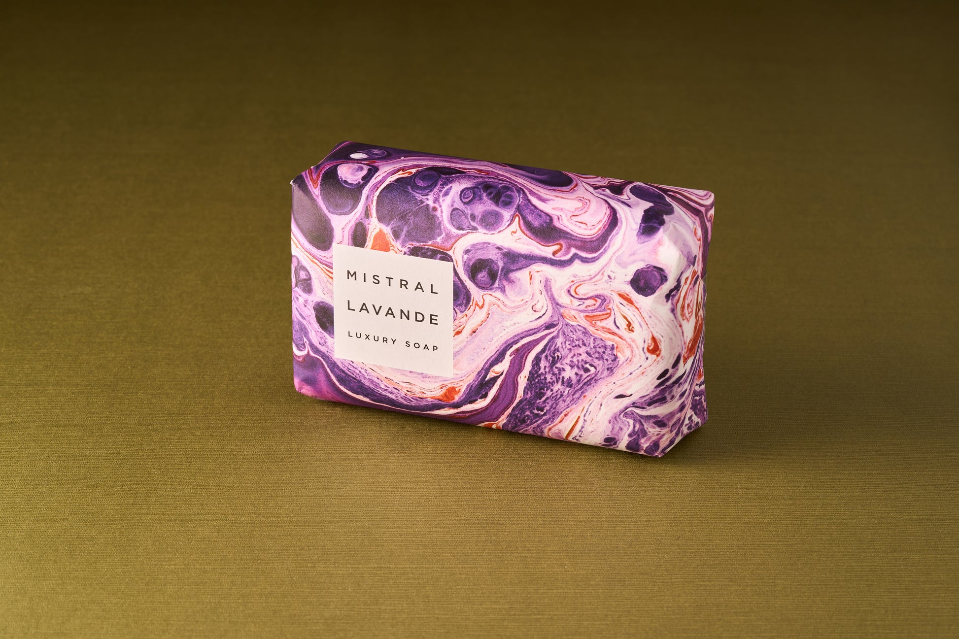 A rectangular Mistral Marbles Lavender Bar Soap, wrapped in marbled purple, white, and orange paper, rests on a gold surface, capturing the luxurious essence of French fragrance.