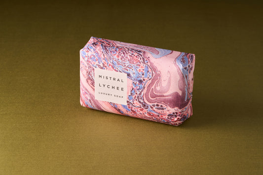 A Mistral Marbles Lychee Rose Bar Soap, a triple-milled French luxury soap, is wrapped in pink, blue, and white marbled paper and placed on a gold textured surface.