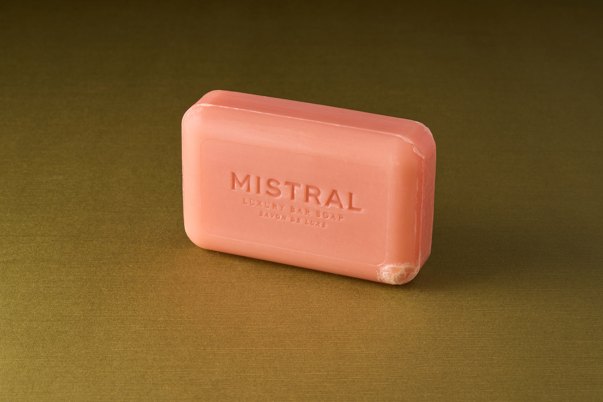 A pink rectangular bar of Mistral Marbles Lychee Rose Bar Soap, a triple-milled soap with a rich French fragrance, rests on a goldish-brown surface.