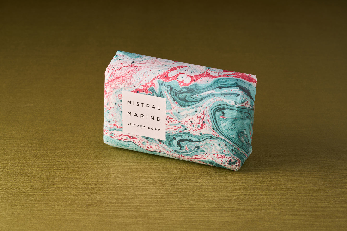 A bar of Mistral Marbles Marine Bar Soap, triple-milled for a creamy lather and wrapped in marbled teal, pink, and white paper, sits on a gold textured surface. Enjoy the essence of French fragrance.