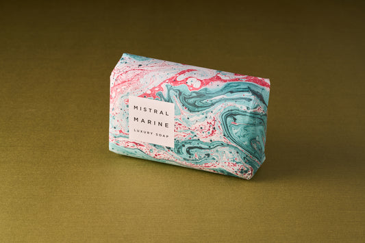 A bar of Mistral Marbles Marine Bar Soap, triple-milled for a creamy lather and wrapped in marbled teal, pink, and white paper, sits on a gold textured surface. Enjoy the essence of French fragrance.