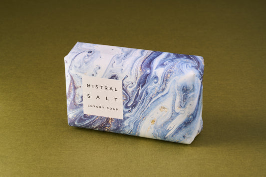 Mistral Marbles Salt Bar Soap in blue-and-white marble packaging with gold accents, resting on an olive-green surface, promising a luxurious, smooth, and fragrant bathing experience.