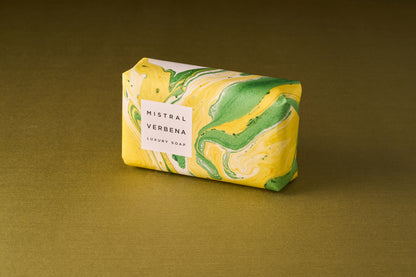 A bar of Mistral Marbles Verbena Bar Soap, cloaked in marbled yellow, green, and white paper, rests on a gold surface—offering an authentic French fragrance experience.