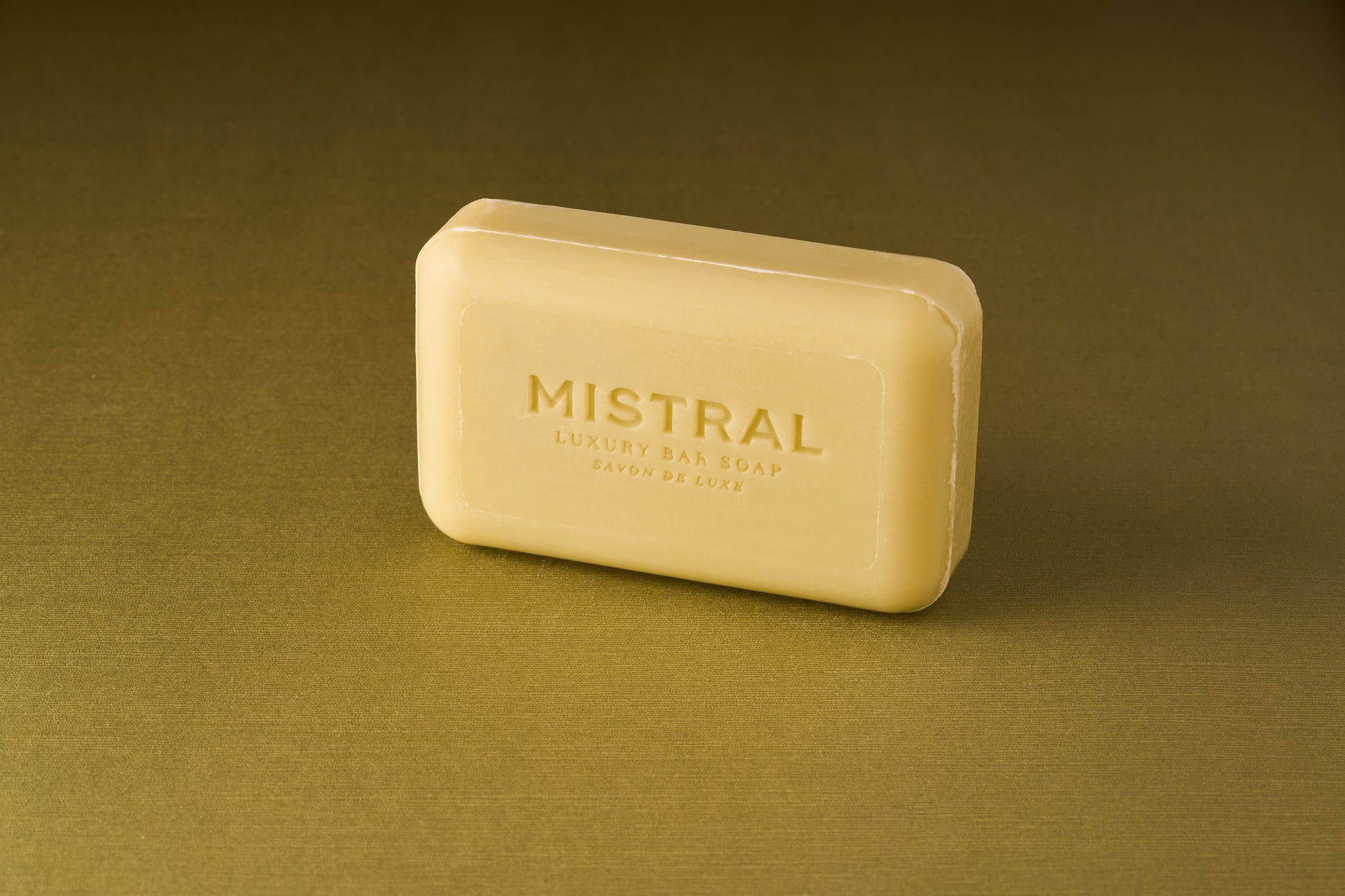 A rectangular yellow bar of Mistral Marbles Verbena Bar Soap stands upright on a gold textured surface with a matching gold background, showcasing its French fragrance and sophisticated charm.