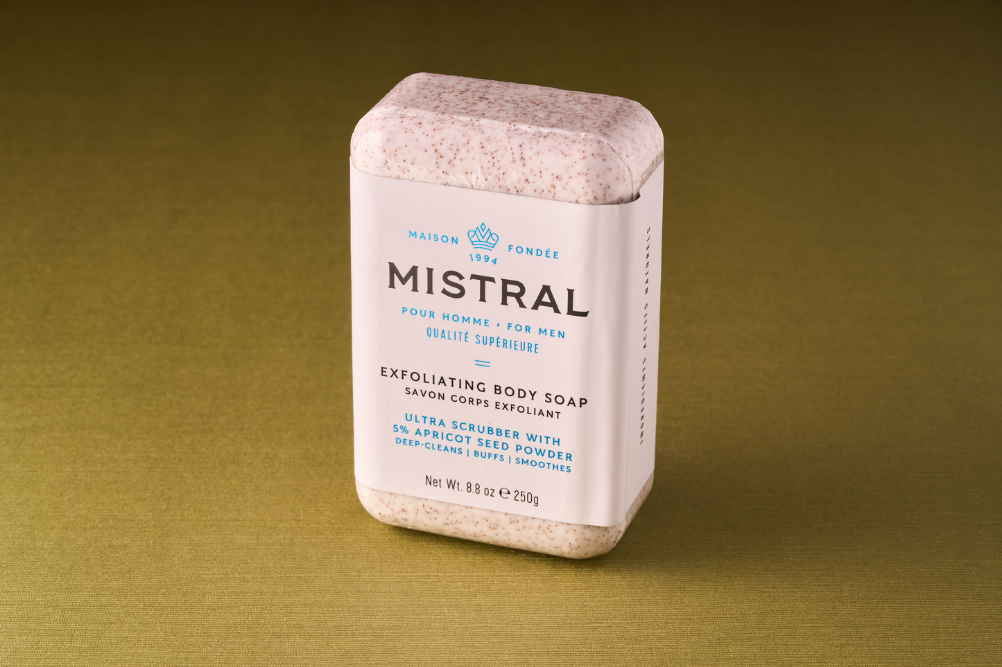 A rectangular bar of Mistral Performance Series Exfoliating Body Soap, wrapped in white packaging with blue text, stands upright on a green-brown surface. The packaging highlights apricot seed powder and a Provence-inspired fragrance.