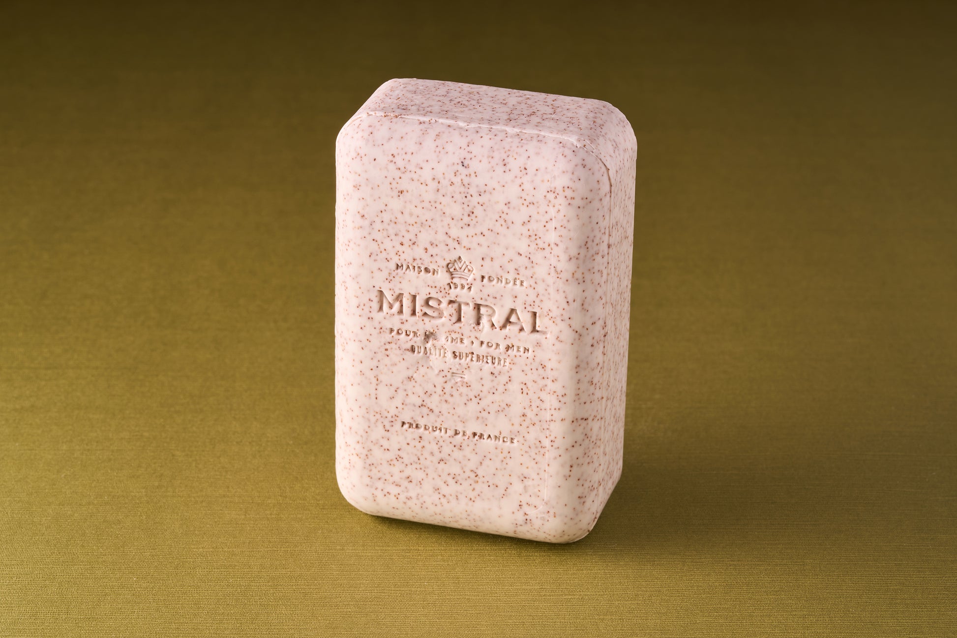 A pink rectangular Mistral Performance Series Exfoliating Body Soap with speckles, embossed text, and a Provence perfume stands upright on a textured olive-green surface.