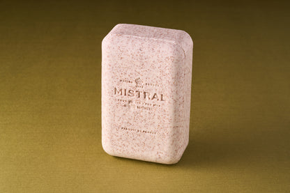 A pink rectangular Mistral Performance Series Exfoliating Body Soap with speckles, embossed text, and a Provence perfume stands upright on a textured olive-green surface.