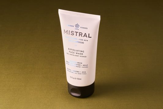 A tube of Mistral Performance Series Exfoliating Face Wash stands upright on an olive green surface. The white tube, accented with black and blue text and a sleek black cap, is inspired by Provence and fine French fragrance.