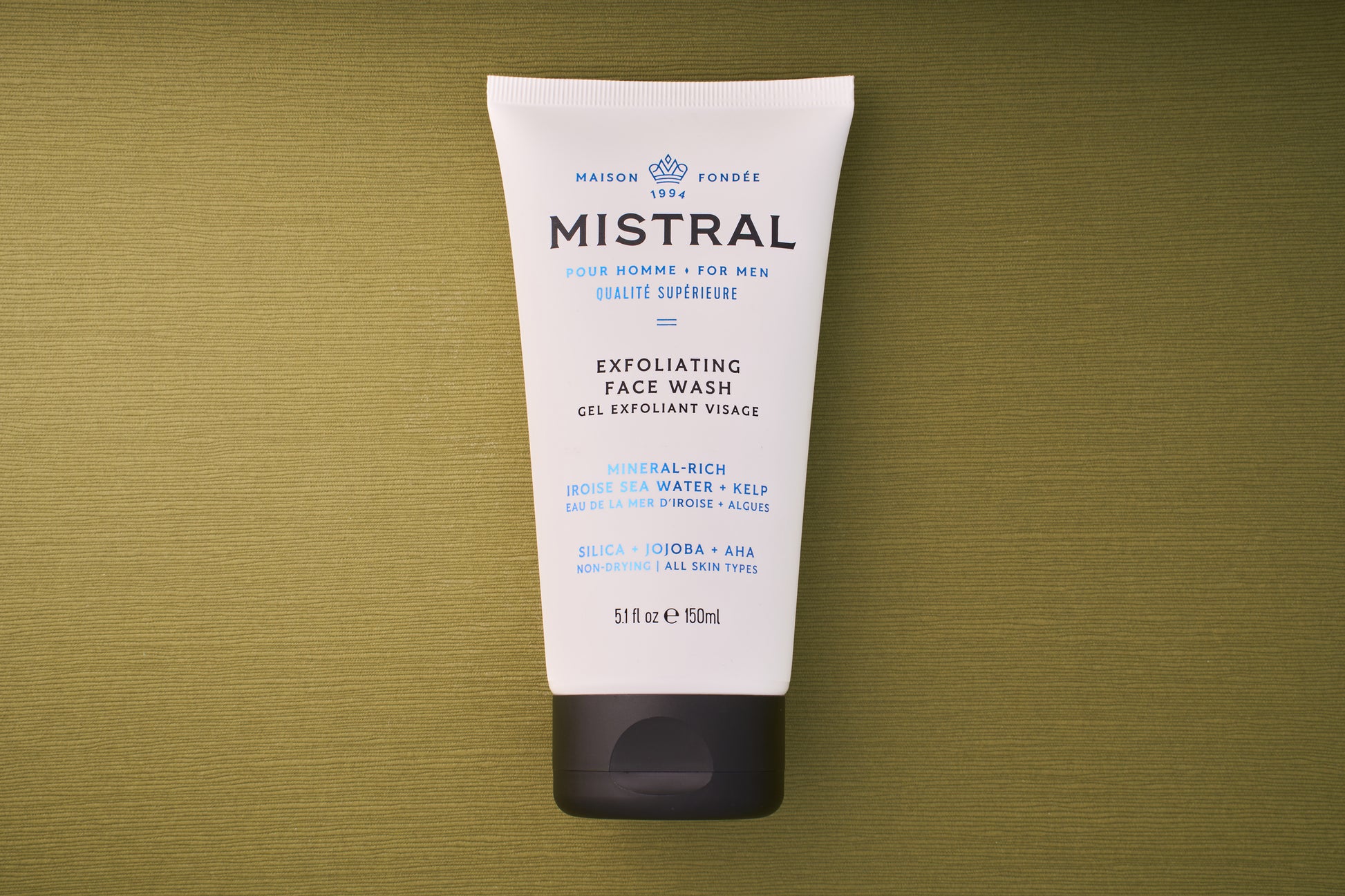 A white tube of Mistral Performance Series Exfoliating Face Wash for men, with blue and black text, sits on a textured olive-green background. Provence-inspired, it offers a French fragrance touch; details and ingredients are shown on the tube.