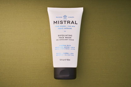 A white tube of Mistral Performance Series Exfoliating Face Wash for men, with blue and black text, sits on a textured olive-green background. Provence-inspired, it offers a French fragrance touch; details and ingredients are shown on the tube.
