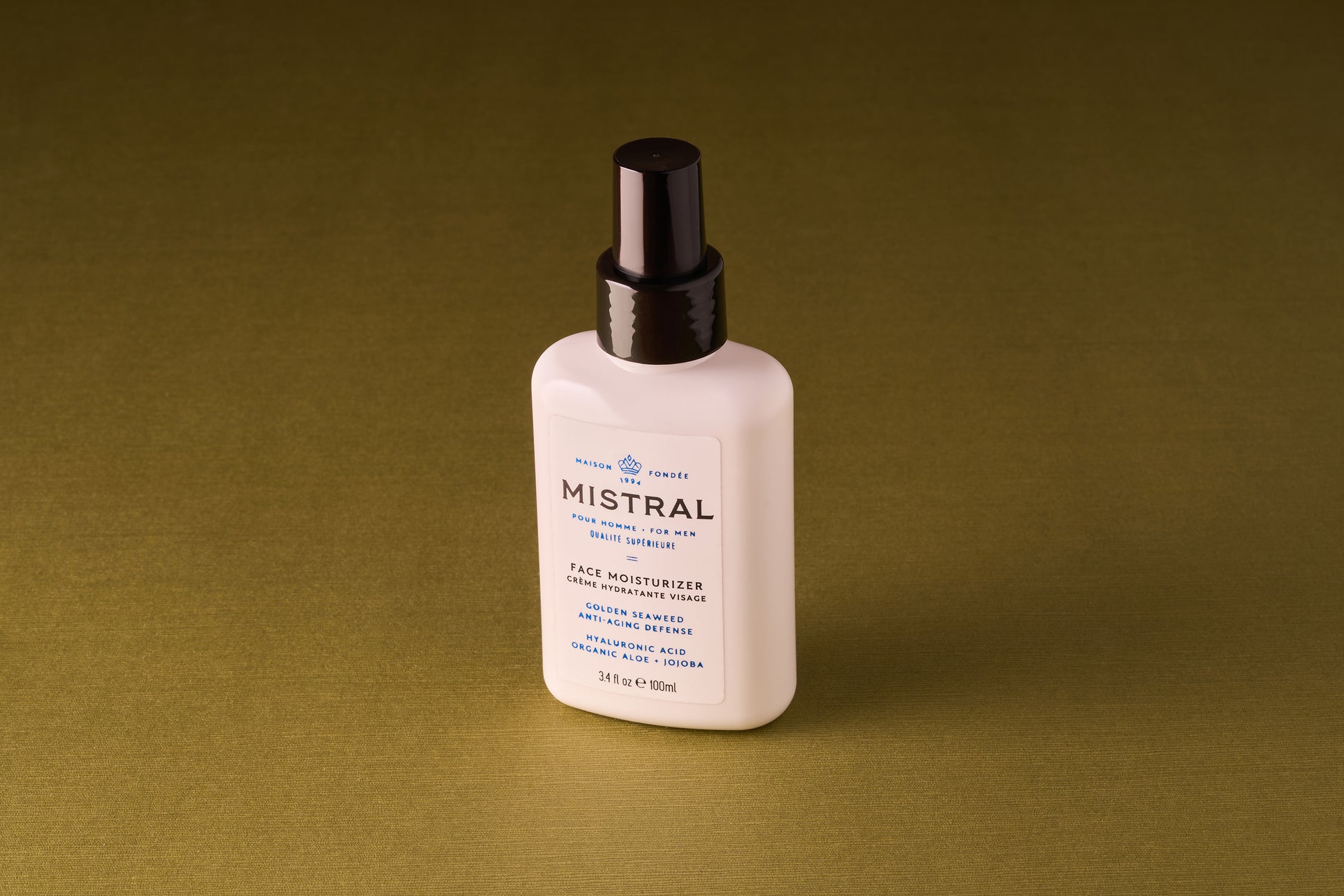 A white bottle of Mistral Performance Series Face Moisturizer with a black pump cap sits against an olive-green background, featuring hydrating actives, organic aloe, and refined French fragrance on its label.