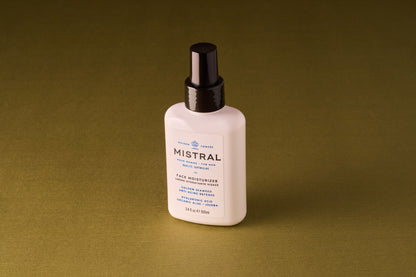 A white bottle of Mistral Performance Series Face Moisturizer with a black pump cap sits against an olive-green background, featuring hydrating actives, organic aloe, and refined French fragrance on its label.