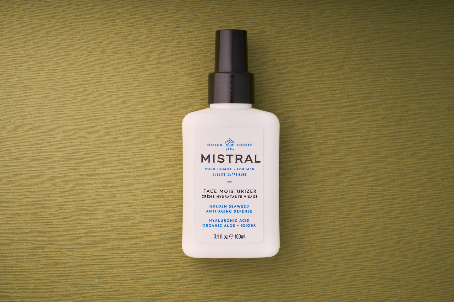 A white 100 ml bottle of Mistral Performance Series Face Moisturizer with a black pump rests on a green textured surface. The label features anti-aging defense with hyaluronic acid, organic aloe, and jojoba, inspired by French fragrance elegance.