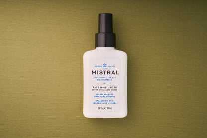 A white 100 ml bottle of Mistral Performance Series Face Moisturizer with a black pump rests on a green textured surface. The label features anti-aging defense with hyaluronic acid, organic aloe, and jojoba, inspired by French fragrance elegance.