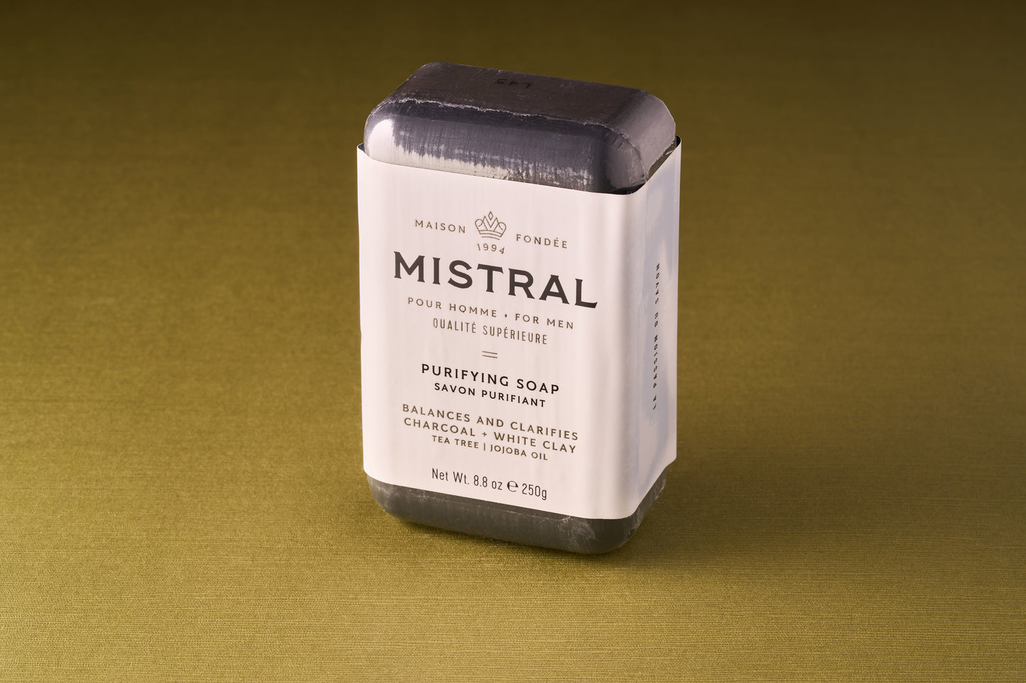 A bar of Mistral Performance Series Purifying Body Soap, wrapped in white paper with black and gold text, rests on an olive green surface. Infused with French fragrance, the soap contains charcoal, white clay, tea tree, and jojoba oil.