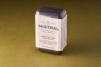 A bar of Mistral Performance Series Purifying Body Soap, wrapped in white paper with black and gold text, rests on an olive green surface. Infused with French fragrance, the soap contains charcoal, white clay, tea tree, and jojoba oil.