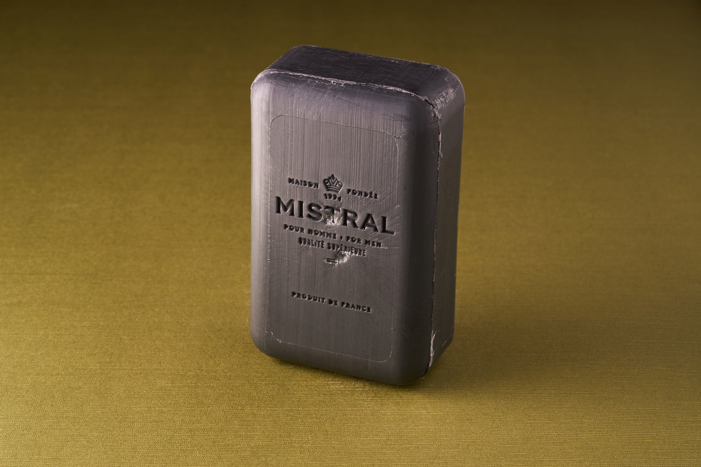 A rectangular bar of Mistral Performance Series Purifying Body Soap stands upright on a textured olive-green surface, its embossed lettering and sleek sheen reflecting the refined style of French-inspired skincare.