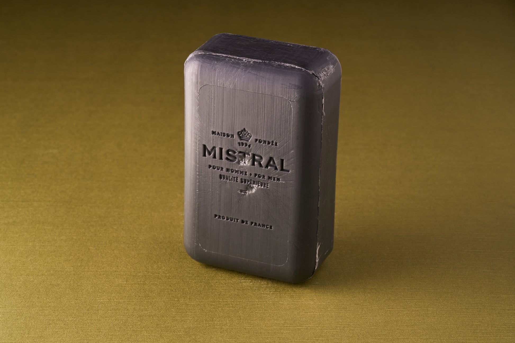 A rectangular bar of Mistral Performance Series Purifying Body Soap stands upright on a textured olive-green surface, its embossed lettering and sleek sheen reflecting the refined style of French-inspired skincare.