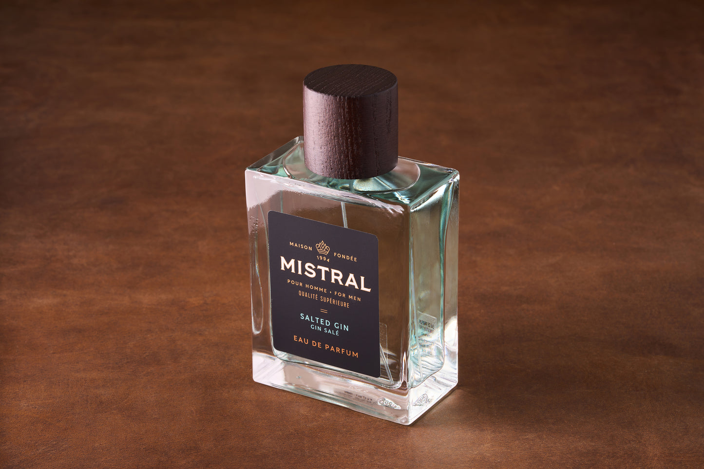 A rectangular glass bottle of Mistral Salted Gin Eau de Parfum 100ml with a dark wooden cap rests on a brown surface; its black label displays elegant white and gold text inspired by Provence perfume.